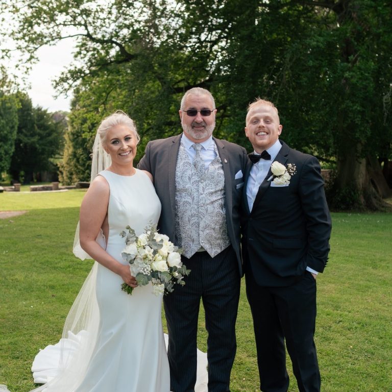 Bride in a white dress, Groom in a dark suit with Celebrant in the middle in and ornate arched structure