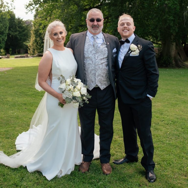 Bride in a white dress, Groom in a dark suit with Celebrant in the middle in and ornate arched structure