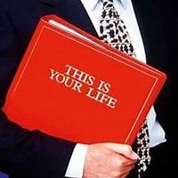 The bust of a man in a dark suit, wearing a white shirt and black and white tie holding a red folder with This is Your Life written in gold