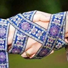 Two hands palms clasped tied together with a blue, gold and pale white ribbon depicting a handfasting ritual